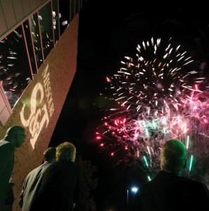 Crowd looks up at the sky watching fireworks. Brick wall of a building is to the left with the Missouri S&amp;T logo projected on to it.