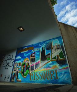 Painted mural with the words "Greetings from Rolla Missouri. Inside the letters of Rolla are painted items representing campus including Joe Miner, St. Pat Statue, residential halls and the Havener Center.
