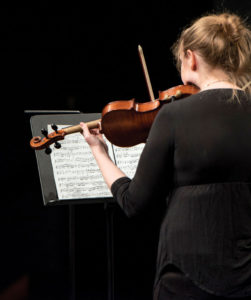 View from behind Sara McCauley dressed in black, standing on stage holding a viola in her left hand and bow in her right. She reads her music sitting on a stand in front of her.